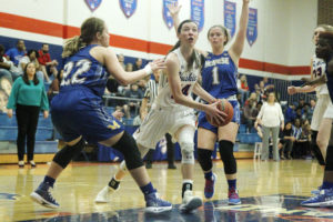 Basketball player in white attempts a layup while two defenders in blue block her path during a women's college game indoors.