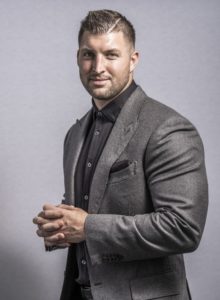 Tim Tebow in a gray suit stands with hands clasped, evoking Spirit of Excellence Gala energy against a light gray background.