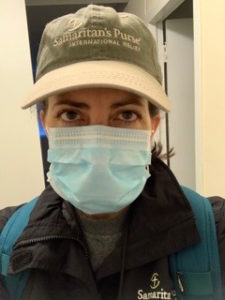 Carrie Hogue in Samaritan’s Purse hat, wearing double masks and jacket, faces camera indoors, likely during a health-related activity.