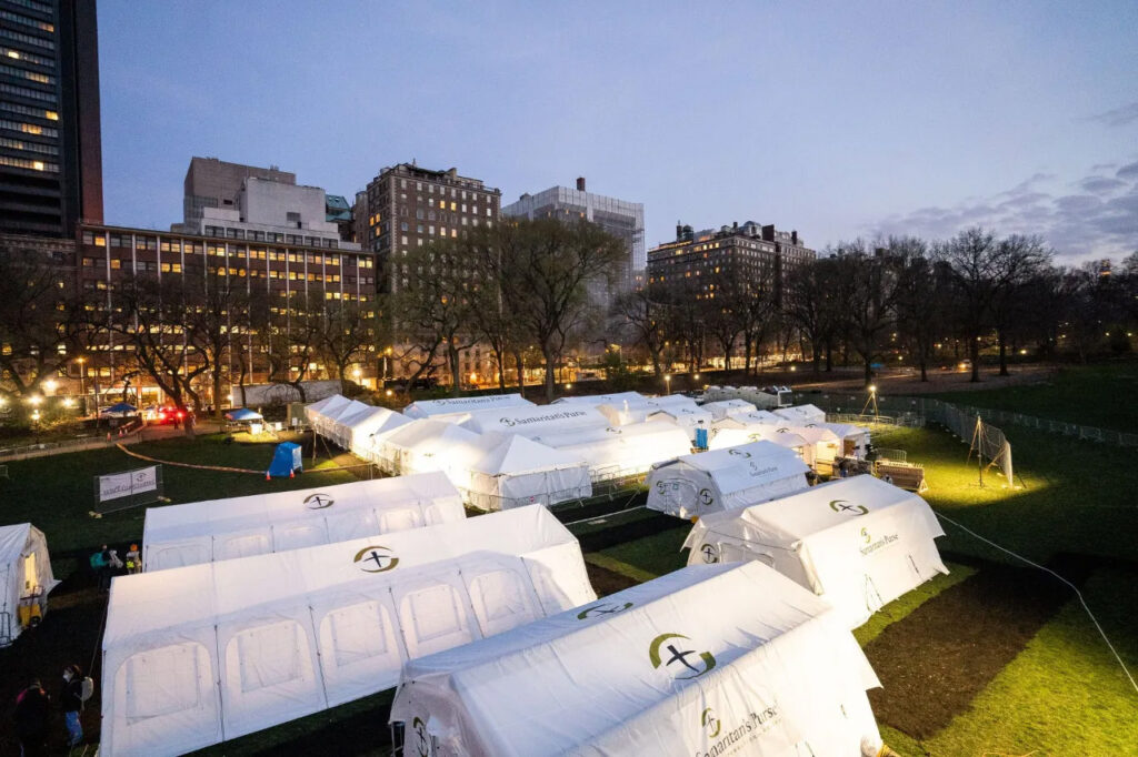 White tents set up in HCU campus green space at dusk, city buildings and trees in background, forming a field hospital.