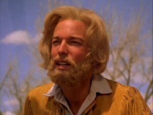 Man with blond hair and beard in a centennial fringe jacket stands outdoors on HCU campus with blue sky and leafless trees.