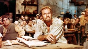 A bearded man in period attire sits with a mug as a crowd observes, evoking a dramatic theater scene during rehearsal.