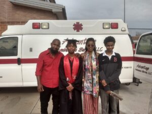 Group of four smiling in front of an HCU ambulance; one wears a graduation cap and gown, celebrating on HCU campus.