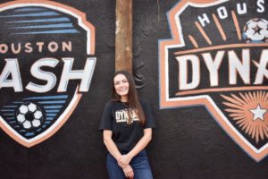 Brielle Eskridge in front of Houston Dash and Dynamo murals on HCU campus, celebrating her accounting degree achievement.