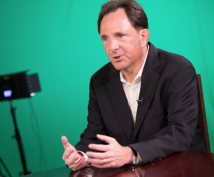 Michael Kraten, in formal attire explains concepts at a table, gesturing as he speaks, against a green screen backdrop.