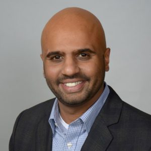 Lijo Joseph, Houston Christian University alumnus, smiles in a dark blazer and checked shirt against a simple backdrop.