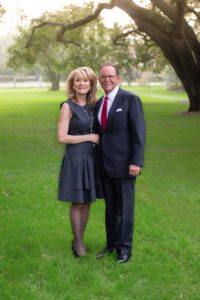 Lisa Simon in a gray dress smiles next to a man in a suit on the HCU campus lawn under a large tree.