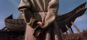 Individual in a robe, hands clasped behind back, reflects on a Bible passage near an unfinished wooden structure on HCU campus.