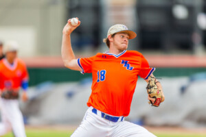 HCU pitcher in orange jersey and tan cap throws on campus field, blurred teammates watching, chasing college baseball dreams.