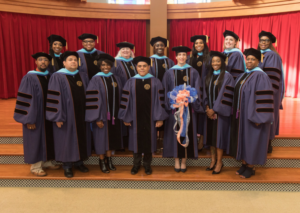 Fifteen graduates in caps and gowns stand on wooden steps indoors by red curtains, celebrating 60 years with joyful expressions.