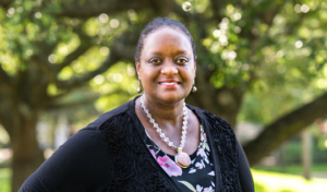 Renata Nero smiles on HCU campus beneath leafy trees, wearing a black cardigan, floral top, and beaded necklace.
