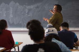Professor explains chemistry equations on a chalkboard as students attentively listen during a university classroom lecture.