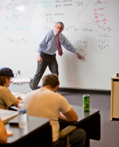 Professor in a tie explains math equations on a whiteboard as university students listen and engage in classroom discussion.
