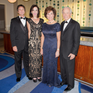 Four guests in formal attire smile by a bar during an indoor Smith Family academic event, engaging in lively conversation.