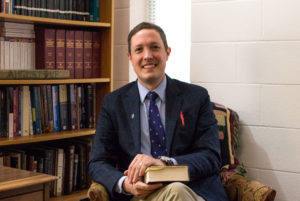 Philip Tallon new dean of the School of Christian Thought in a suit, seated with book, surrounded by bookshelves in academic office.