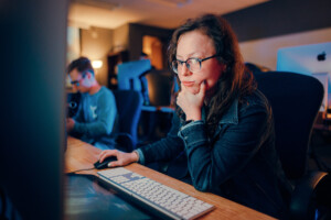 Student with glasses focuses on computer, composing prompts in a collaborative academic office environment with peers nearby.