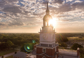 Belin Tower on HCU campus stands tall with sunlight streaming behind, framed by trees and scattered clouds in the sky.