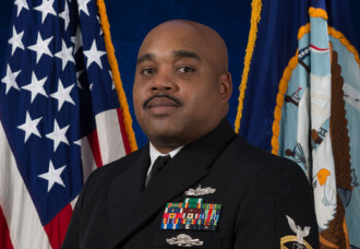 Navy Chief Darryl Smith in uniform stands before American and Navy flags, embodying Christian leadership values.