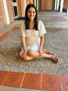Young woman in a white dress smiles beside President’s Award bricks on HCU campus, marking her Senior Year Experience.