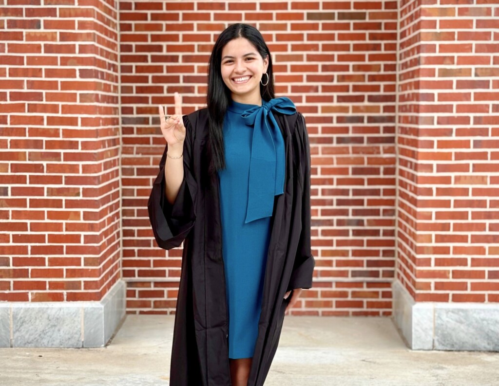 Andrea Ramirez, in a graduation gown, smiles by a brick wall on the HCU campus celebrating her senior year.