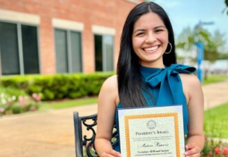 Andrea Ramirez smiles while holding a framed President’s Award certificate outdoors on the HCU campus during her senior year.