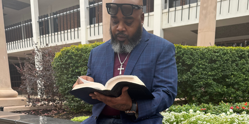 Navy Chief Darryl Smith in blue blazer and sunglasses reads outside near garden and building on HCU campus.