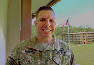 A smiling U.S. Army veteran, wounded warrior, stands outside on HCU campus with a flag and building visible in the background.
