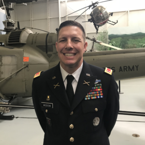 U.S. Army officer in dress uniform, wounded warrior, smiles while standing before a military helicopter in an academic hangar.