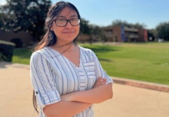 Smiling student with glasses and long hair stands outside on HCU campus, pursuing her dreams with Belin Tower in the background.