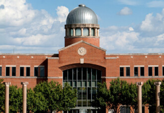 HCU campus view of Archie W. Dunham College of Business, red brick building with central dome, framed by trees and columns.