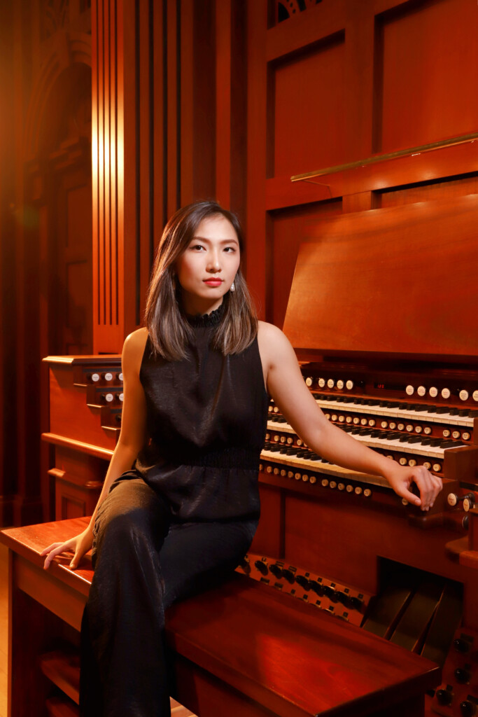 Valentina Huang in a black outfit plays the grand pipe organ during an organ recital, hand poised on the keys in concert hall.