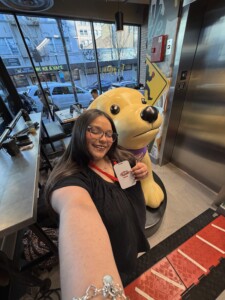 Graduate snapping a selfie beside a large yellow dog statue in a campus cafe, holding a cup with red logo.