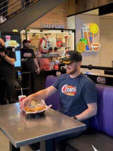 A student in a Canes t-shirt and cap smiles at a table of chicken tenders with friends during a campus celebration.