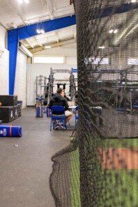 Kinesiology graduate and another person converse on blue chairs in a gym with exercise equipment, viewed through a net.