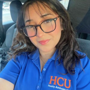 First-generation nursing student with glasses and wavy hair, smiling in her car, wearing blue Husky Ambassador shirt.