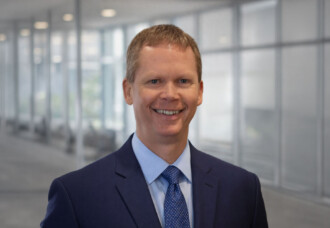 Kevin Hartless, VP of Online Adult & Graduate Studies, smiles in a glass-walled academic office corridor.