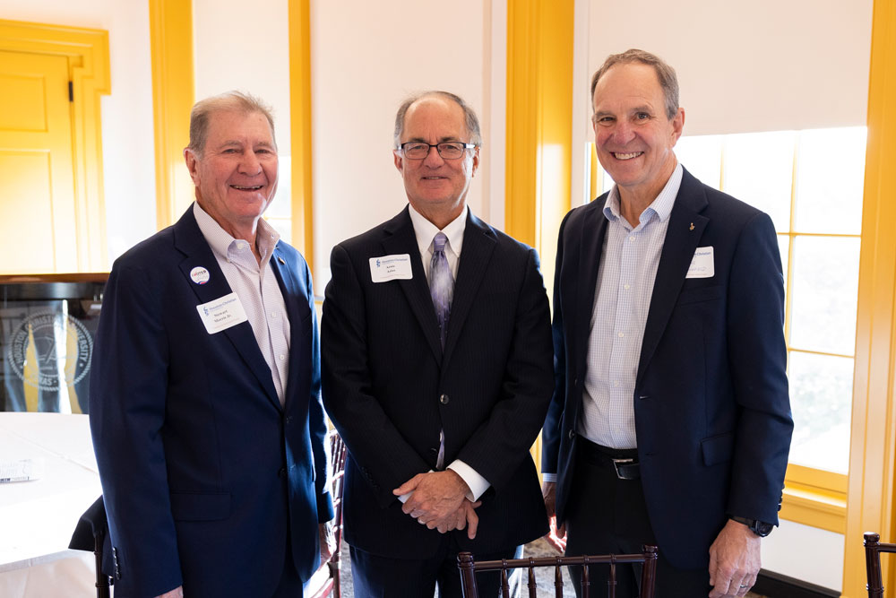 Three men in suits smiling at a formal campus event, yellow window frames hinting at HCU Student Center backdrop.
