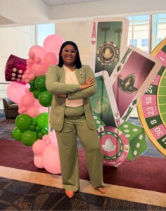 Smiling woman in a green suit stands with arms crossed at indoor casino-themed event with festive balloon decorations.