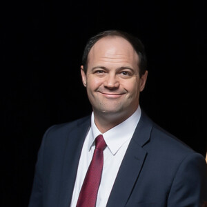 Smiling man in a suit and red tie stands before a plain black background, dressed for professional or academic presentation.