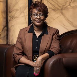 Professor in glasses and brown blazer sits on a leather chair, smiling with hands folded in her lap during classroom discussion.