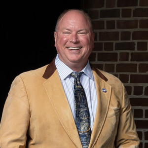 Man in tan blazer with brown collar, striped shirt, and patterned tie stands by HCU campus brick wall, smiling confidently.