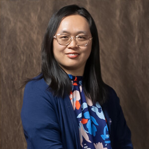Faculty member with long dark hair, glasses, blue floral top, and blazer stands before a plain brown academic backdrop indoors.