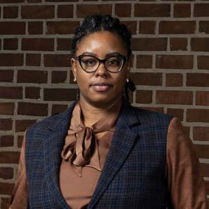 A woman wearing glasses and a brown outfit stands by a brick wall on the HCU campus, facing the camera directly.