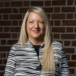 Student with long blonde hair in a striped top smiles by a brick wall on the HCU campus, showcasing campus life.