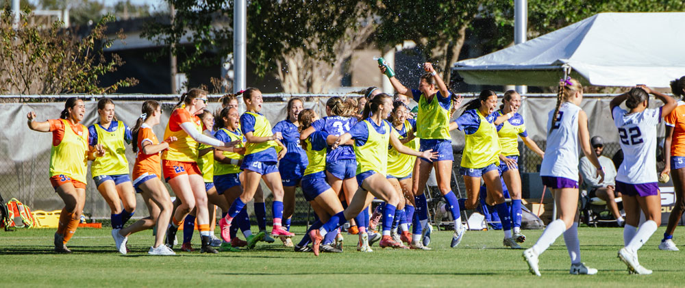 HCU Huskies soccer players celebrate on the field, some soaked with water, united in excitement to make history.