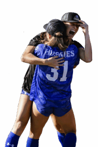 Two female soccer players wearing hats hug and smile, one in a blue Huskies 31 jersey, celebrating a historic win on HCU campus.