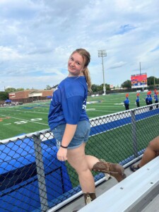 Sophia poses in the stands at the HCU campus football field with the marching band on the sidelines.