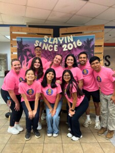 Students in matching pink shirts pose for a group photo in front of an HCU Slayin Since 2016 backdrop at Houston Christian.