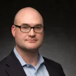 Man with glasses and bald head in dark blazer and light blue shirt at Apologetics Day event, posing against dark backdrop.
