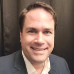 Man in suit jacket and white shirt smiles against a dark backdrop at Apologetics Day, highlighting academic engagement.